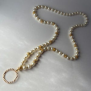 May include: A long lanyard necklace featuring a strand of white faux pearls interspersed with small gold-toned spacers and clear crystal accents. The necklace terminates in a circular gold-toned metal ring with a textured, diamond-cut surface.