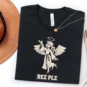 Könnte beinhalten: Ein schwarzes Kurzarm-T-Shirt mit einer cremefarbenen Grafik eines Engels mit Heiligenschein, Flügeln und einem Gewand. Unter dem Engel steht der Text REZ PLZ in einer fetten Blockschrift. Das T-Shirt hat einen Rundhalsausschnitt und ein weißes Etikett.