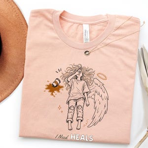 May include: A light peach-colored t-shirt featuring a black line art graphic of a winged figure with a halo, covering their eyes with one hand. A spilled coffee cup is nearby. The text "I NEED HEALS" is printed in white at the bottom of the shirt.