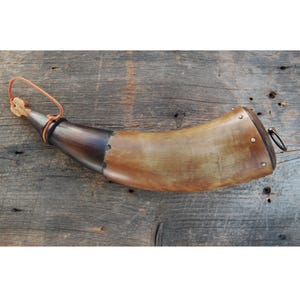 Puede incluir: Un cuerno de pólvora curvo hecho de cuerno animal natural con una punta marrón oscura y un cuerpo de color beige claro. El extremo ancho está tapado con un tapón de madera asegurado por remaches metálicos y cuenta con una pequeña anilla.