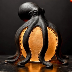 Può includere: Una scultura di polpo color grigio antracite con una superficie ruvida e otto tentacoli ricurvi che avvolgono una fonte di luce sferica centrale color ambra, creando un'illuminazione calda dall'interno del corpo della creatura.