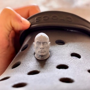 May include: A dark brown foam clog shoe featuring a small, light gray, three-dimensional sculpted head of a person attached as a decorative charm in one of the circular ventilation holes. The word Crocs is embossed on the heel strap.