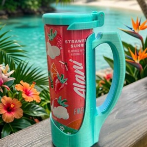 May include: A teal 3D-printed plastic can holder with a handle and a hinged lid, securely holding a red Alani Strawberry Sunrise energy drink can. The can features graphics of strawberries and orange slices with the text Alani Energy.