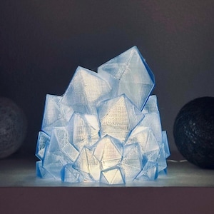 May include: A translucent, light blue, geometric crystal-shaped lamp with a faceted surface. The structure is composed of multiple angular, pyramid-like segments that emit a soft, internal glow, creating a cluster of illuminated, crystalline shapes.