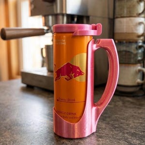 May include: An orange Red Bull energy drink can held within a custom-fitted, 3D-printed pink plastic mug holder. The holder features a sturdy side handle and a matching pink lid that secures the top of the can, creating a mug-like drinking vessel.