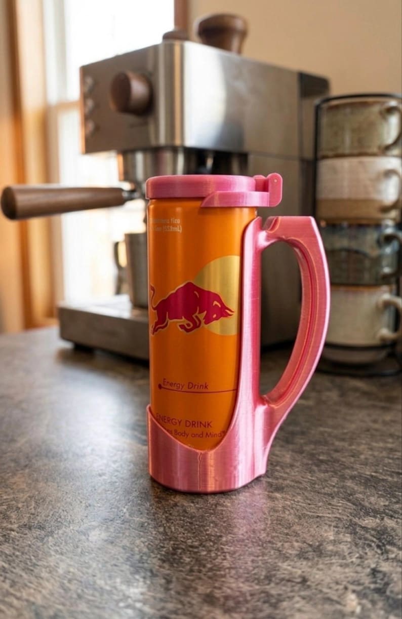 May include: An orange Red Bull energy drink can held within a custom-fitted, 3D-printed pink plastic mug holder. The holder features a sturdy side handle and a matching pink lid that secures the top of the can, creating a mug-like drinking vessel.