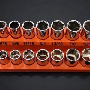 Op de afbeelding: Een set zilverkleurige metalen dopsleutels gerangschikt in twee rijen op een oranje plastic opbergtray. De bovenste rij toont de maten 7/16, 1/2, 9/16, 5/8, 11/16, 3/4, 13/16, 7/8, 15/16 en 1 inch in zwarte opdruk op de tray.