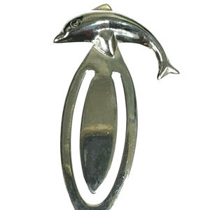 Puede incluir: Marcapáginas de metal plateado con diseño de delfín. El delfín está en la parte superior, con el cuerpo curvado. El marcapáginas tiene forma ovalada con una ranura para insertar páginas.