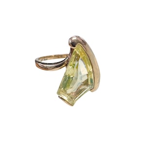 Könnte beinhalten: Ein goldfarbener Ring mit einem großen, facettierten, blassgelben Edelstein in Trapezform. Das Metallband wölbt sich nach oben und rahmt eine Seite des Steins ein, wodurch ein asymmetrisches Design entsteht, das die lichtreflektierenden Facetten betont.
