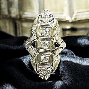 Pu&ograve; includere: Un anello d'argento con un design geometrico che presenta cinque diamanti rotondi disposti in fila. L'anello ha un design delicato e intricato con un'estetica vintage.