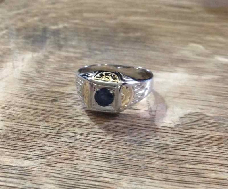 May include: A silver and gold ring with a blue gemstone set in the center. The ring has intricate details and a textured band.