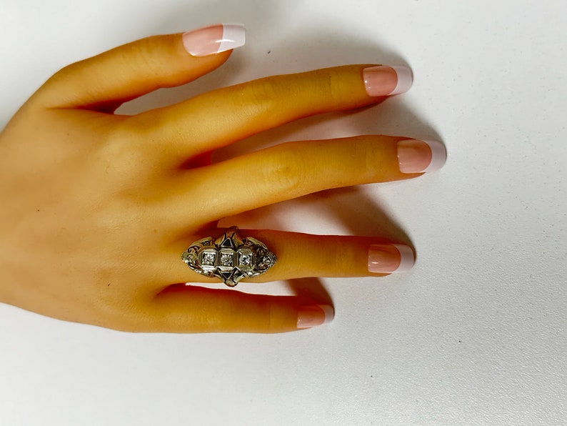 Pu&ograve; includere: Un anello d'oro con un gruppo di pietre chiare incastonate in un design decorativo. L'anello &egrave; su una mano con smalto bianco.
