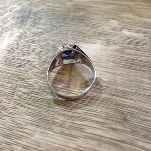 May include: A silver ring with a blue gemstone set in a square bezel. The ring is on a wooden surface.