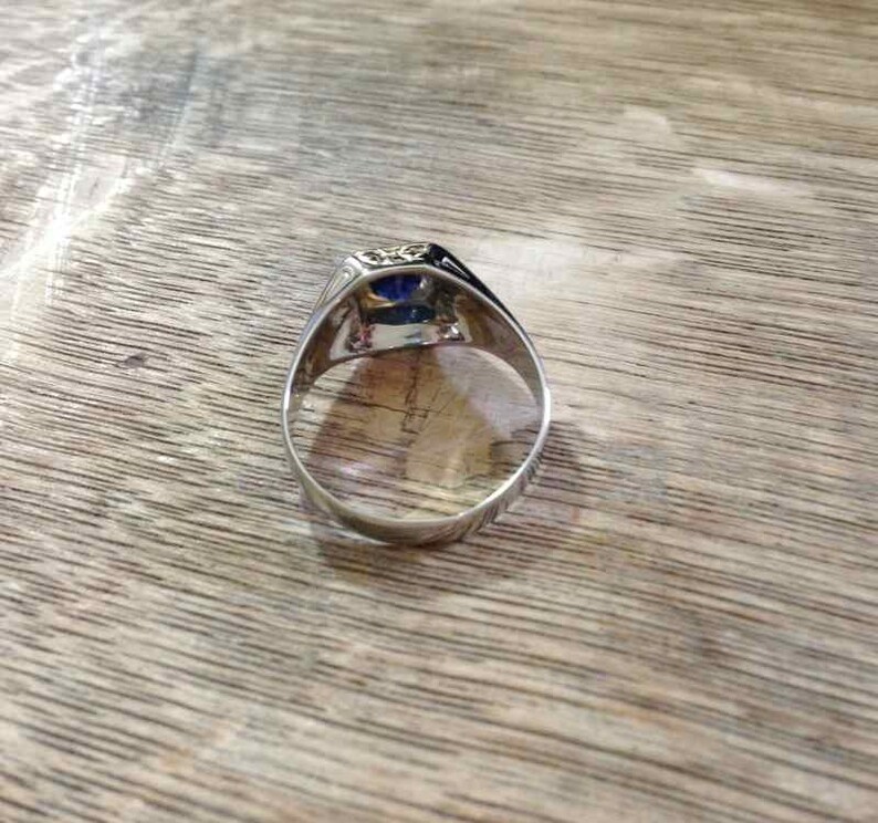 May include: A silver ring with a blue gemstone set in a square bezel. The ring is on a wooden surface.