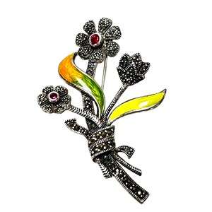 May include: A silver brooch with a bouquet of flowers made of marcasite and enamel. The flowers are yellow, green, and red. The brooch is tied with a bow.