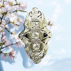 Pu&ograve; includere: Un anello d'argento con un delicato design a filigrana e tre diamanti rotondi incastonati in una forma quadrata. L'anello ha due pietre nere su entrambi i lati dei diamanti.