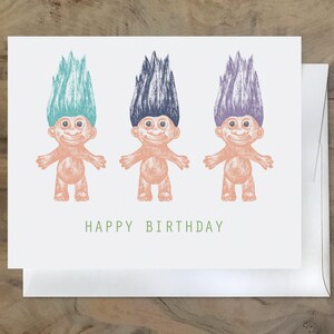 May include: A greeting card with three cartoon trolls with different colored hair. The trolls are smiling and have their arms outstretched. The card says "Happy Birthday" in green text.