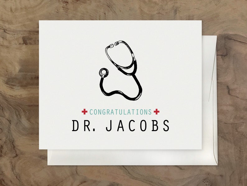 May include: A white card with a black and white illustration of a stethoscope. The card has red plus signs and the text "CONGRATULATIONS" and "DR. JACOBS" in teal.
