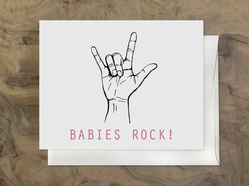 Puede incluir: Una tarjeta blanca con una ilustraci&oacute;n en blanco y negro de una mano haciendo el gesto de rock and roll. El texto "BABIES ROCK!" est&aacute; impreso en rosa debajo de la ilustraci&oacute;n.