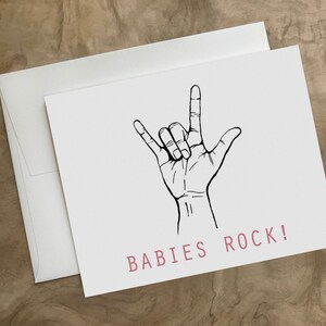Puede incluir: Una tarjeta blanca con un dibujo en l&iacute;nea negra de una mano haciendo el gesto de rock and roll. El texto "BABIES ROCK!" est&aacute; impreso en rosa en la parte inferior de la tarjeta.