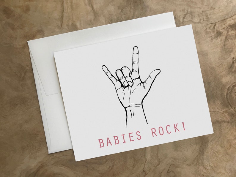 Puede incluir: Una tarjeta blanca con un dibujo en l&iacute;nea negra de una mano haciendo el gesto de rock and roll. El texto "BABIES ROCK!" est&aacute; impreso en rosa en la parte inferior de la tarjeta.