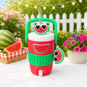 May include: A red and white beverage cooler with a green handle and a spigot at the base. The cooler features three watermelon slice decorations with heart-shaped sunglasses, a clear straw, and a green textured band around the middle section.