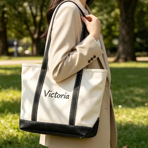 Puede incluir: Bolso tote color crema con detalles negros y el nombre "Victoria" impreso en la parte delantera. El bolso tiene asas negras y una base negra. Una persona con una blazer beige sostiene el bolso.