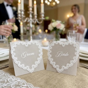 Puede incluir: Dos tarjetas de mesa de cartulina beige con forma de corazón, cada una con un borde de encaje blanco y texto en cursiva blanca. Una tarjeta muestra la palabra Groom y la otra la palabra Bride, ambas de pie sobre una superficie de tela.