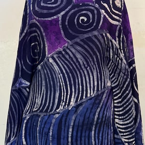 May include: A lightweight fabric wrap featuring a batik-style design with deep purple and navy blue tones. The pattern consists of large circular spirals at the top and sections of vertical, curved lines throughout the rest of the textile surface.