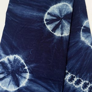 May include: A deep navy blue fabric featuring a tie-dye pattern with large, circular white motifs. The design includes several distinct, radiating sunburst-like circles and a smaller, linear row of circular patterns along the right edge.