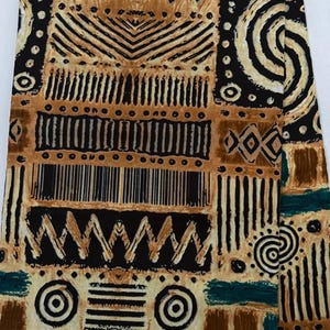 May include: Folded fabric with a tribal pattern in black, brown, and tan, with teal accents. The design includes geometric shapes and lines. The fabric has a textured appearance. The words "TRIBAL WAX 703246" are printed on the bottom.