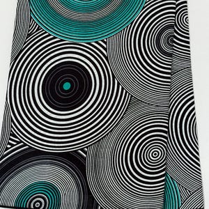 May include: Fabric featuring a bold, repeating pattern of concentric circles in black, white, and teal. The design creates a striking visual effect. The words "GUARANTEED AFRICAN" are printed along the edge of the fabric.