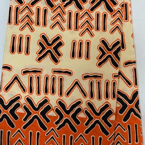 May include: Cream-coloured fabric with a repeating geometric pattern in black and orange. The design features stylized shapes resembling arrows, crosses, and vertical lines. The fabric has a black border with the text "VERITABLE BLOCK PR".