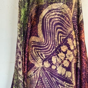 May include: A long, flowing garment featuring a batik-style pattern in shades of deep purple, lime green, and burnt orange. The fabric displays intricate, hand-drawn wavy lines, geometric shapes, and a mottled, textured effect across the surface.