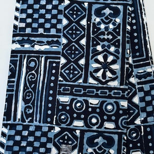 Op de afbeelding: Een stof met een geometrisch patroon in donker marineblauw en lichtblauw met herhalende vierkanten, cirkels en bloemmotieven. Het textiel bevat een klein label aan de onderrand met de tekst INDIGO BY KENNYBEE en het woord ORIGINAL.
