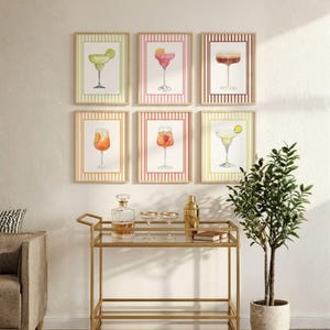 May include: Six framed wall art prints featuring watercolor illustrations of various cocktails, including margaritas and spritzes, arranged in two rows of three. Each print is encased in a light wood frame with a striped border matching the drink color.