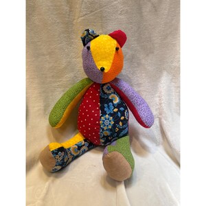 Pode incluir: Um urso de peluche feito de vários retalhos de tecido em cores vibrantes, incluindo amarelo, laranja, vermelho, verde, roxo e azul. O urso apresenta um padrão floral no tronco e nas orelhas, bolinhas no peito e olhos de contas pretas.