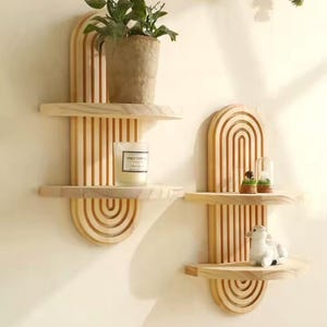 Puede incluir: Dos estantes de pared de madera con acabado natural. Cada estante tiene un diseño superior e inferior arqueado con listones verticales. Uno contiene una planta en maceta y una vela, el otro pequeños objetos decorativos.