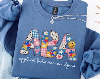 May include: An indigo blue crewneck sweatshirt featuring the letters ABA in a patchwork design with puzzle pieces, flowers, hearts, and a smiley face. Below the letters, the text reads applied behavior analysis in white cursive script.