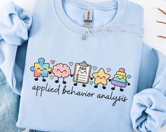 May include: A light blue crewneck sweatshirt featuring a graphic of a puzzle piece, a brain, a clipboard labeled ABA, a star, and a stacking toy, all with smiling faces. The text "applied behavior analysis" is printed below the graphic.