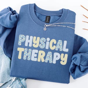 May include: An indigo blue crewneck sweatshirt featuring the text "PHYSICAL THERAPY" in a bold, rounded font. The letters are filled with a pattern of light blue daisies, yellow gingham checks, and light blue vertical stripes.