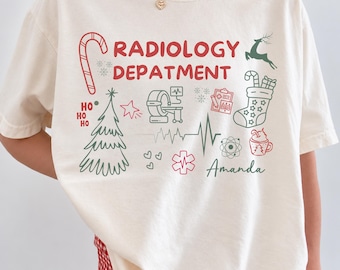 May include: An off-white t-shirt featuring the red text RADIOLOGY DEPATMENT and various green and red holiday-themed icons including a candy cane, Christmas tree, medical scanner, EKG line, stocking, and the name Amanda in script.