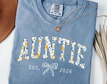 May include: A blue short-sleeved t-shirt with the word AUNTIE printed in a white-outlined floral font. Below the text is a white line-art bow graphic with the text EST. 2026. The shirt features a Comfort Colors brand label.