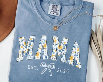 May include: A blue short-sleeve t-shirt featuring the word MAMA in large block letters filled with a yellow and blue floral pattern. Below the text, the shirt displays the white text EST. 2026 with a simple line-drawn bow graphic in the center.