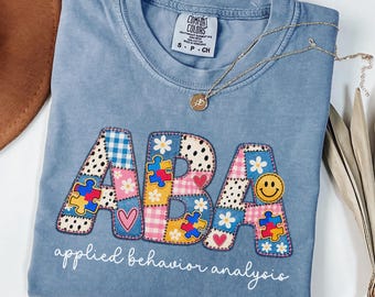May include: A blue short-sleeved t-shirt with a crew neckline featuring the letters ABA in a patchwork design of blue, pink, and yellow patterns, including puzzle pieces, hearts, and smiley faces, with the text applied behavior analysis below.