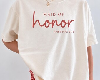 May include: An off-white short-sleeve t-shirt featuring the red text "MAID OF honor OBVIOUSLY." printed on the chest. The shirt has a crew neckline and a relaxed fit, paired with red and white gingham patterned shorts.