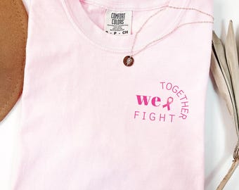 Puede incluir: Una camiseta de manga corta de color rosa claro con cuello redondo. En el pecho lleva el texto "TOGETHER WE FIGHT" en rosa oscuro en semicírculo, con un lazo rosa que sustituye a la letra "I" en la palabra "WE". Etiqueta: COMFORT COLORS.