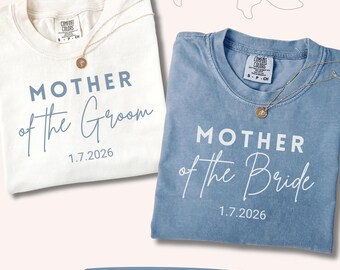 May include: An ivory t-shirt with blue text reading MOTHER of the Groom 1.7.2026 and a blue t-shirt with white text reading MOTHER of the Bride 1.7.2026. Both shirts feature a Comfort Colors label and a small gold pendant necklace.
