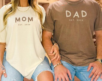 May include: A cream-colored t-shirt with the text MOM EST. 2026 and a brown t-shirt with the text DAD EST. 2026. Both shirts feature short sleeves and a crew neckline, worn with blue denim jeans that have distressed knee cutouts.