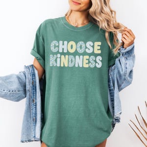 Puede incluir: Una camiseta de manga corta color verde bosque con el texto CHOOSE KINDNESS en una fuente redondeada y negrita. Las letras est&aacute;n rellenas con un patr&oacute;n repetitivo de peque&ntilde;as flores amarillas, margaritas azul claro y rayas verticales.